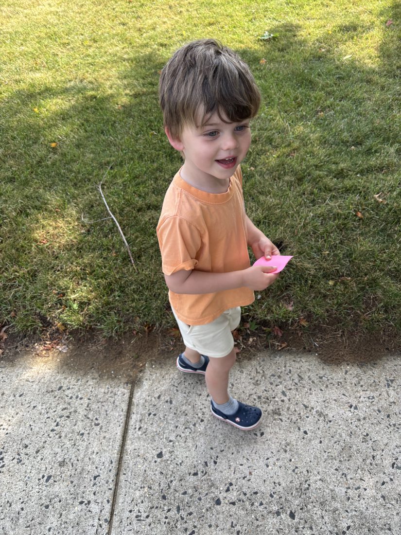 A young boy in an orange shirt standing on the sidewalk during a mailbox hunt, holding his tally note