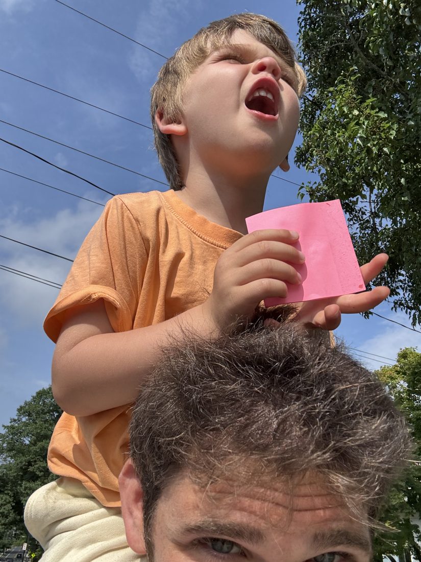 A boy riding on his dad's shoulders outside, laughing, holding his mailbox-hunt tally note