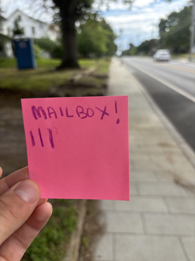 A hand holding a pink sticky note reading MAILBOX with three tally marks, a real mailbox and street behind it